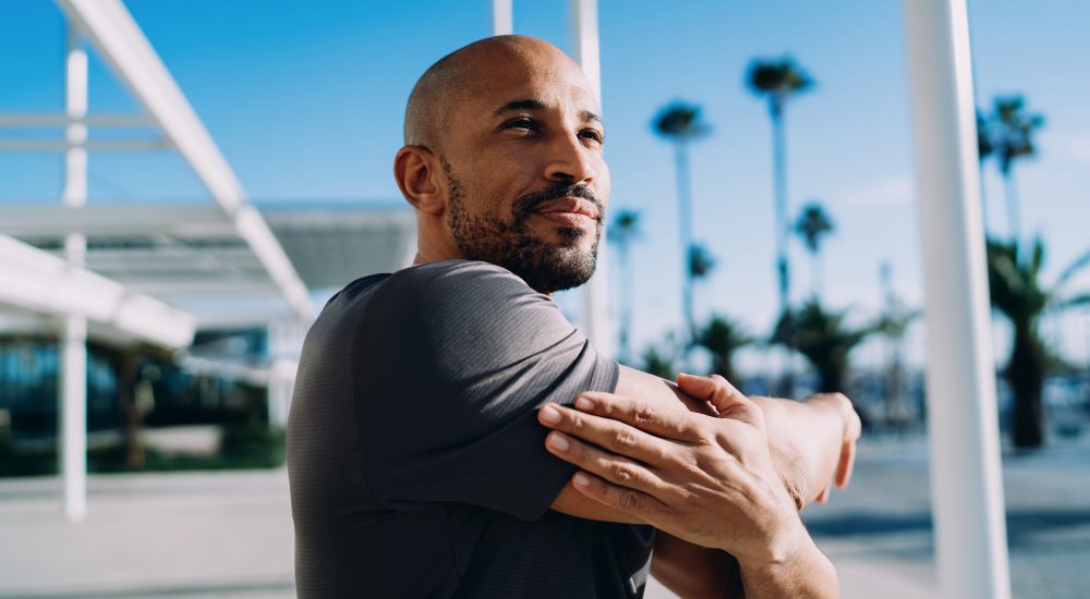 Low angle portrait of Black man stretching shoulder against blue sky and palm trees, focus on fitness and wellness, authentic representation, mindfulness, serenity, tropical city vibes.