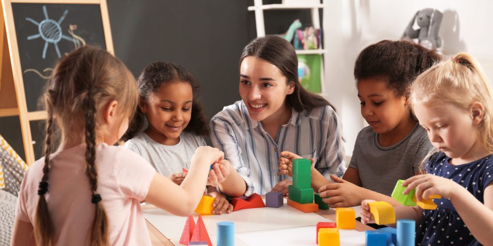 Cute little children and nursery teacher playing with building blocks in kindergarten. Indoor activity