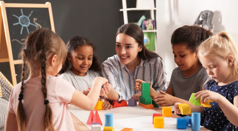 Cute little children and nursery teacher playing with building blocks in kindergarten. Indoor activity