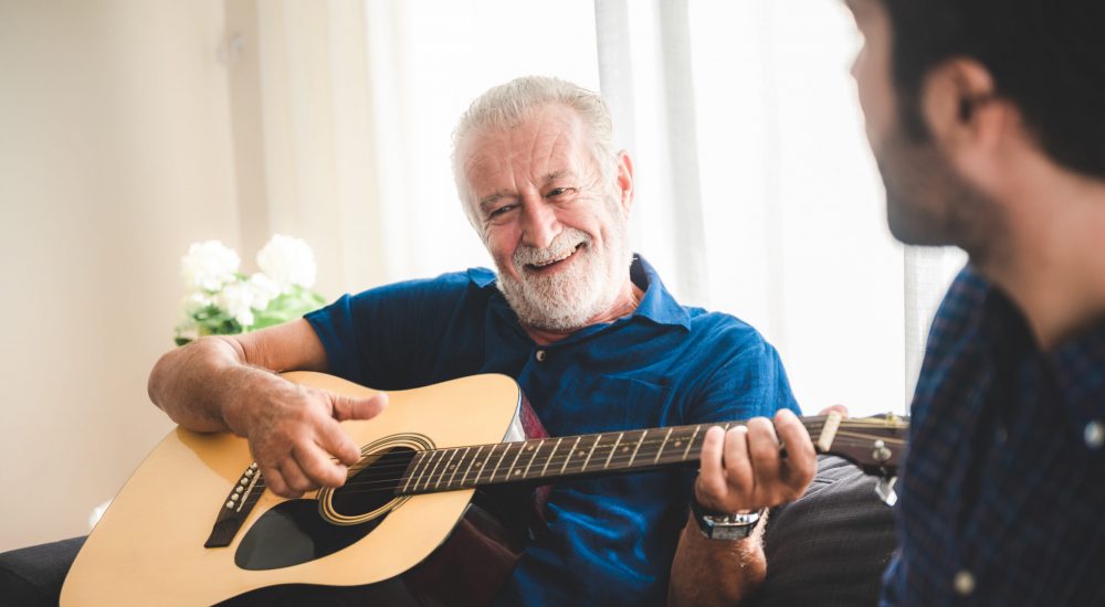 happy adult son and senior father playing guitar on sofa at home relaxing and having happy time with family