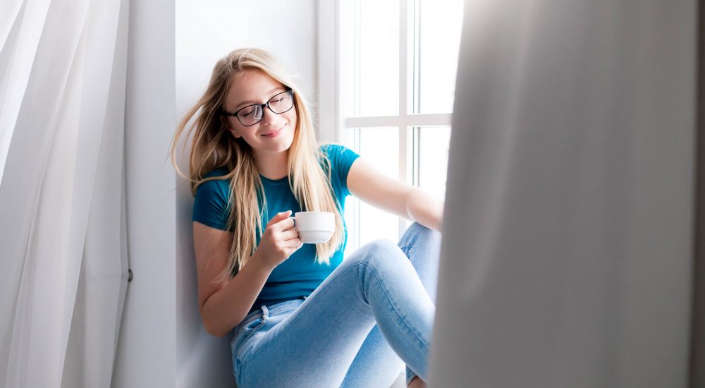 Young woman relaxing with cup of coffee on cozy windowsill at home. Girl enjoying fresh air and wind from window with curtains. Self isolation on safe place, social distance during quarantine.
