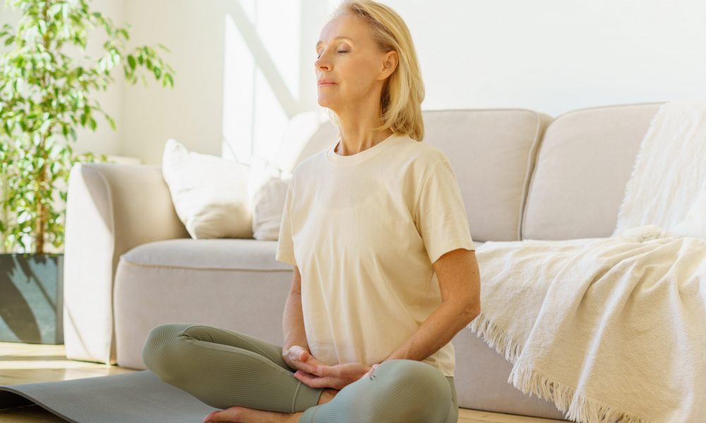 Peaceful senior woman in lotus position meditation with closed eyes at home while sitting on yoga mat on floor, full length. Calm elderly lady practicing meditation techniques and yoga indoors