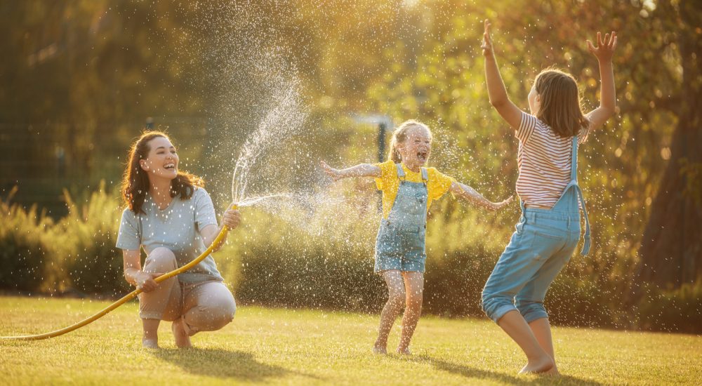 Happy family playing in backyard. Mother sprinkling her kids in hot summer day.