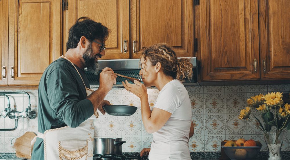 One happy real couple at home enjoying life together and relationship. Man giving food to woman to taste. Male people cooking for his wife. Male and female enjoying leisure activity indoor in kitchen
