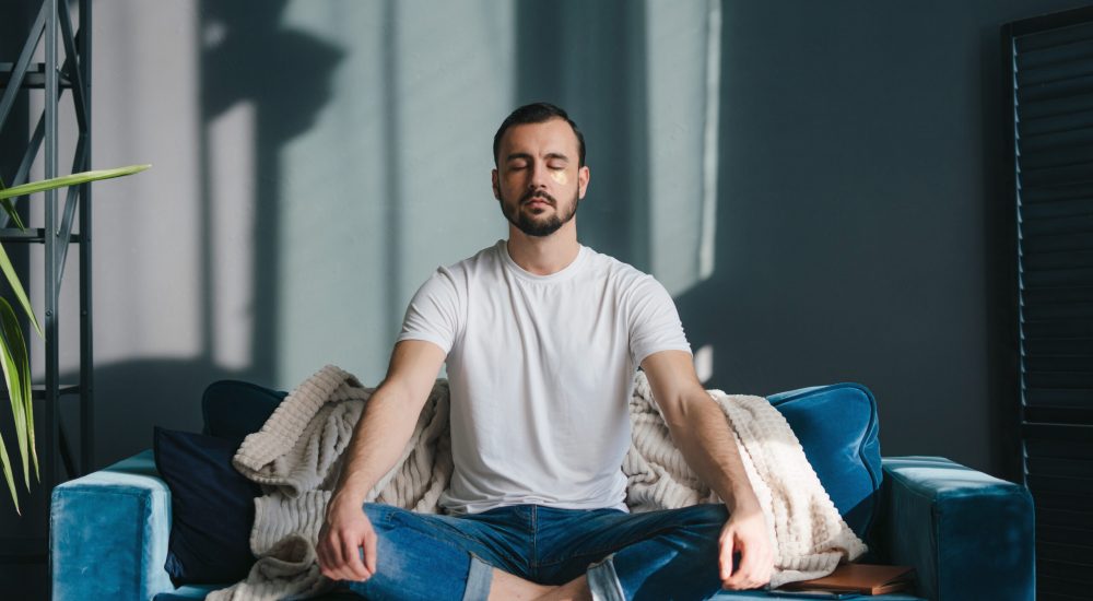 Focused man meditating in lotus pose breathing deep and slowly wearing casual clothes sitting on comfortable coach relaxing at home. Feeling free from stress.