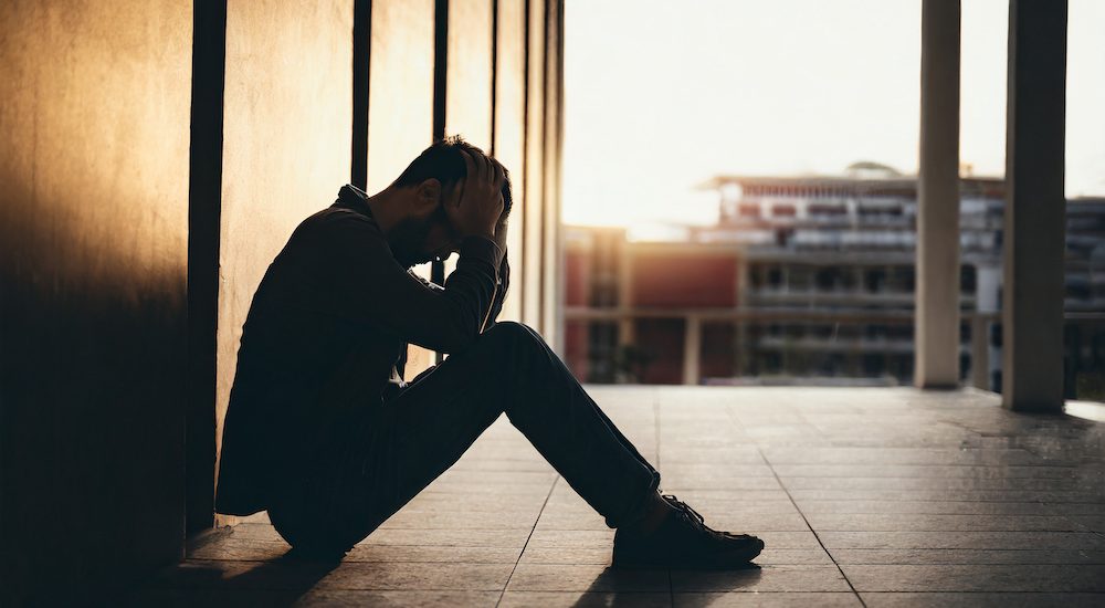 Silhouette of despondent man, head in hands, seated on residence walkway