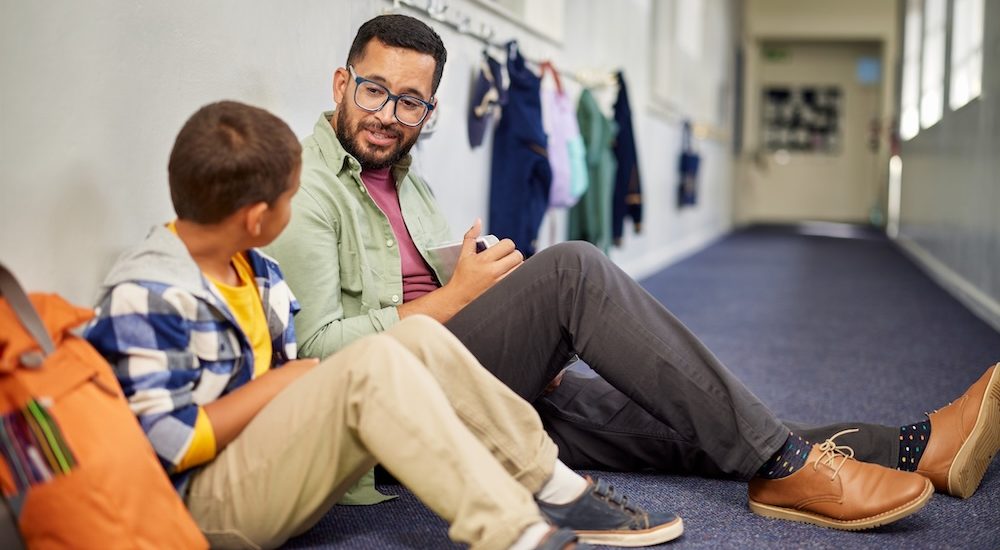 Young male psychologist speaking to depressed boy in school while sitting on the floor in school corridor. Man working with a child while discussing about learning problem and bullying. Young and friendly teacher speaking to boy in school hallway.