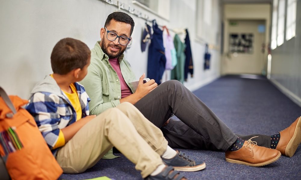 Young male psychologist speaking to depressed boy in school while sitting on the floor in school corridor. Man working with a child while discussing about learning problem and bullying. Young and friendly teacher speaking to boy in school hallway.