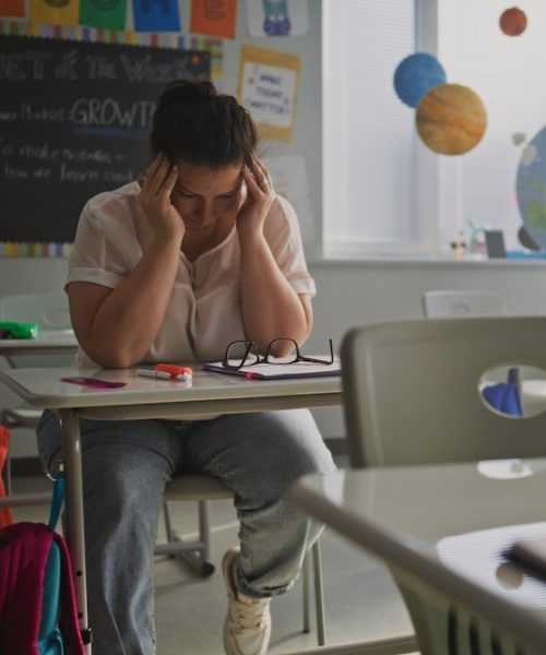 Exhausted Female Teacher Rubbing her Temples, Sitting Alone at Desk in Empty Classroom, Having Rest After Lesson. Woman Feeling Stress, Burnout and Emotional Struggle, Working in Modern Primary School